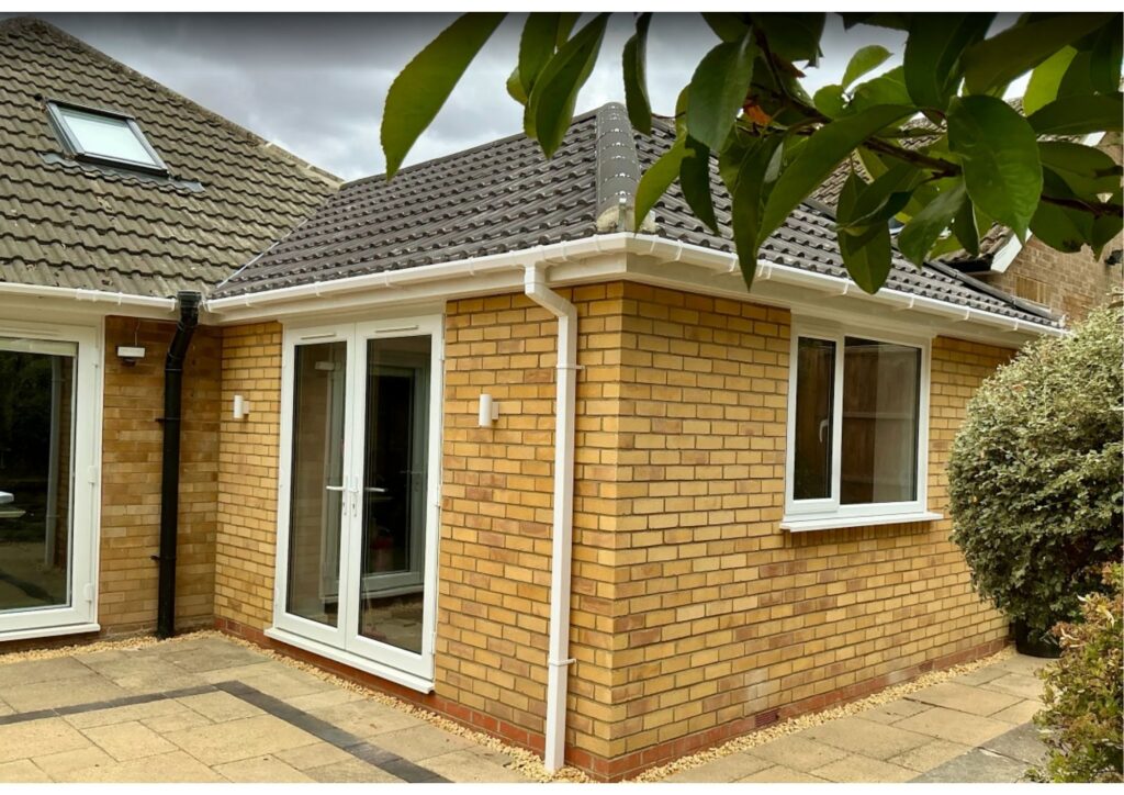Corner of a brick house with white-framed glass doors and windows, brown tiled roof, and a tree branch in the foreground.